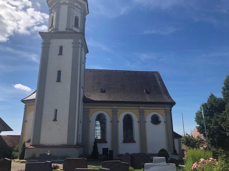 Weißes Kirchengebäude mit Turm unter blauem Himmel und wenigen Wolken.