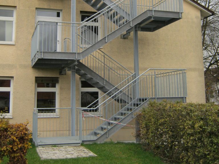 Graue Außentreppe an einem mehrstöckigen Gebäude, umgeben von Büschen.