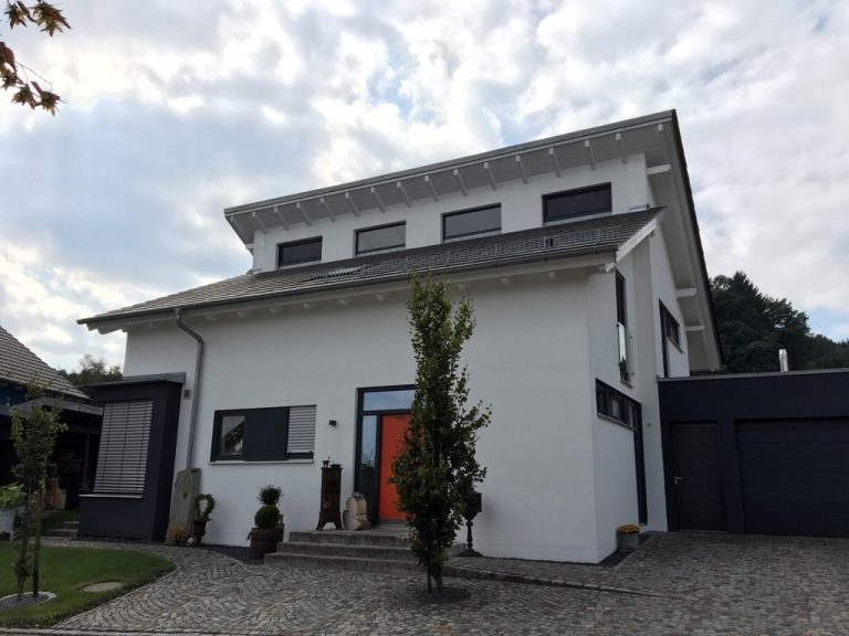 Modernes weißes Haus mit orangefarbener Eingangstür und umgeben von Bäumen.