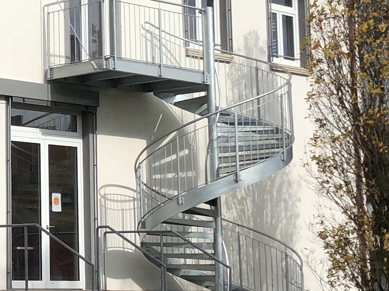 Eisenwendeltreppe an einem Gebäude mit Fenster und gelben Bäumen im Hintergrund.