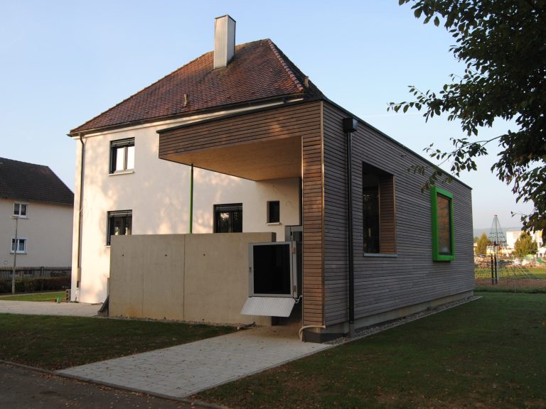 Modernes, weißes Wohnhaus mit Garage und grüner Umgebung unter bewölktem Himmel.