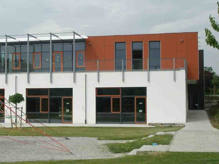 Moderne Gebäude mit großer Fensterfront, orangefarbenem Akzent und Grünflächen davor.