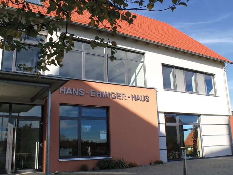 Eingang zum Hans-Ehinger-Haus mit moderner Architektur und großen Fenstern.