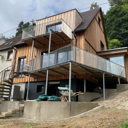 Modernes Holzhaus mit Balkon, unter dem eine Terrasse und Werkzeuge sichtbar sind.