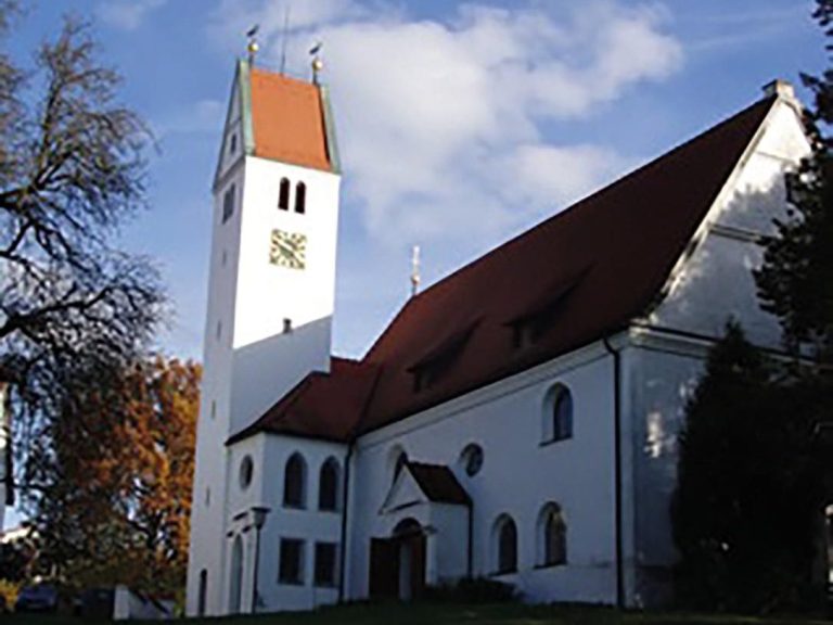 Weißes Gebäude mit rotem Dach und hoher Kirchturm, umgeben von Bäumen und blauem Himmel.