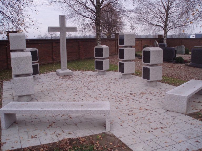 Weiße Betonbänke und kreuzförmiges Denkmal in einem ruhigen, begrenzten Raum.